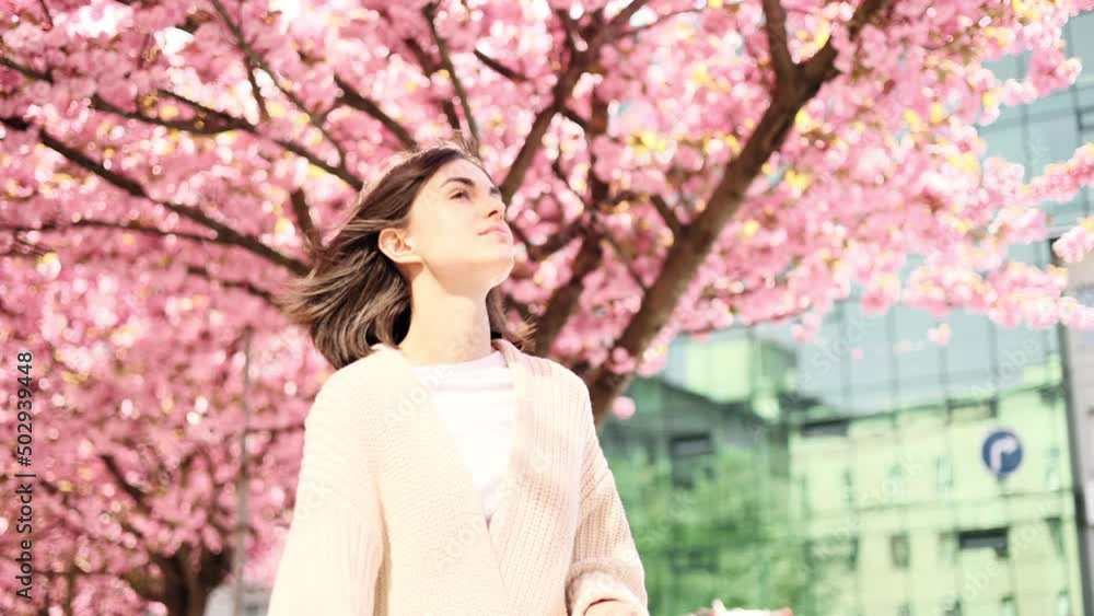 Portrait of a cute young woman while walking down the city street with sakura flowers in the background. Beautiful woman looking ahead outside. Portrait of a cute young woman while walking down the city street with sakura flowers in the background. Beautiful woman looking ahead outside.