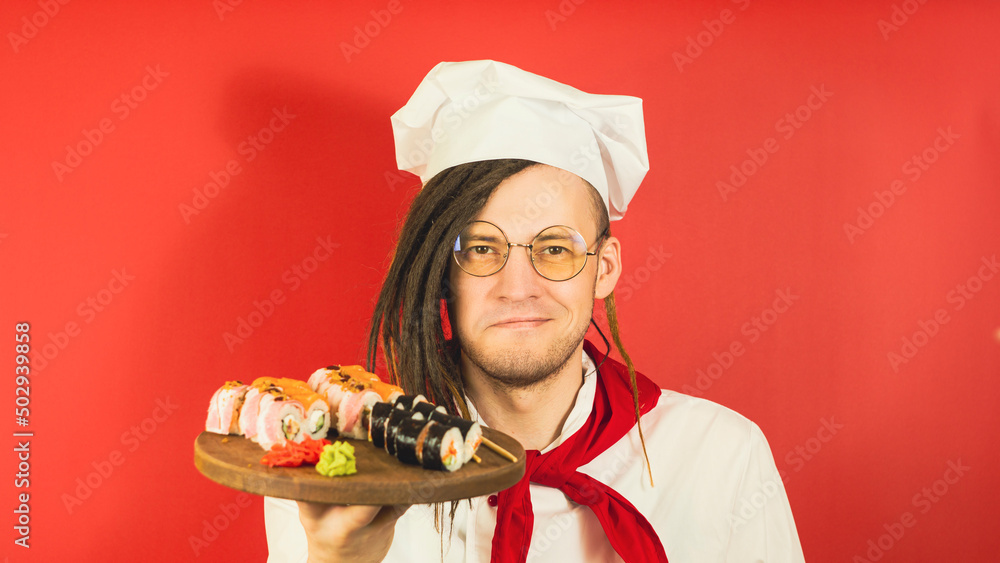 Young man in glasses dressed as chef holding wooden board with sushi rolls. Male cook with appetizing sushi rolls on red background. Young man in glasses dressed as chef holding wooden board with sushi rolls. Male cook with appetizing sushi rolls on red background.