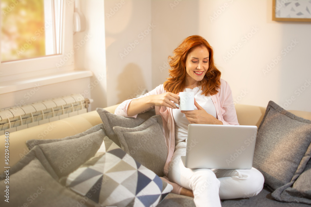 Beautiful ginger woman holding cup of coffee and reading news on laptop while relaxing on sofa at home in the morning. Beautiful ginger woman holding cup of coffee and reading news on laptop while relaxing on sofa at home in the morning.