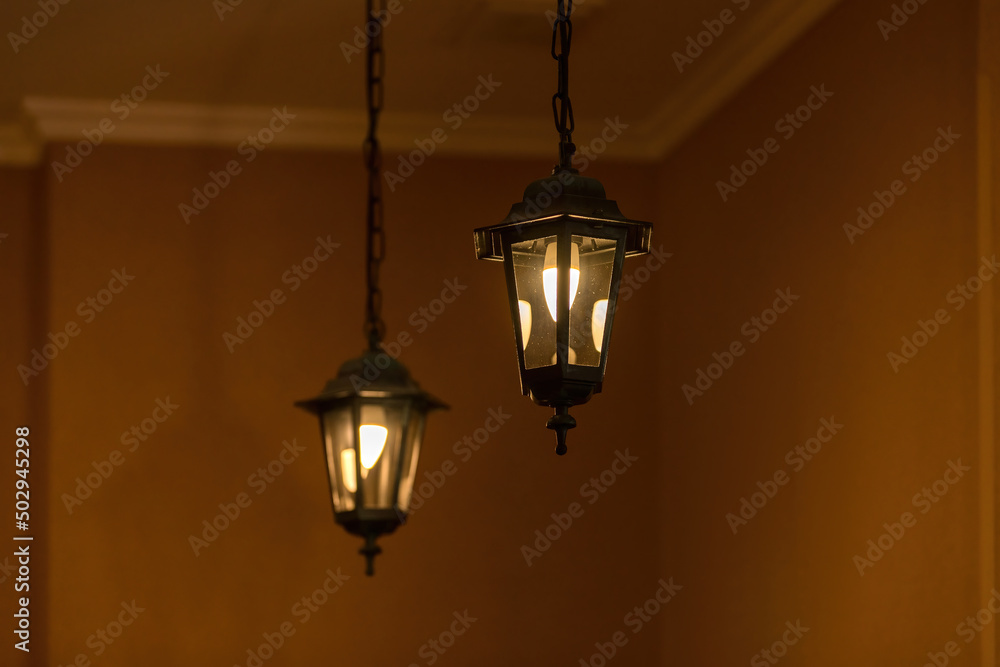 Two vintage electric lighting lanterns hang on chains and glow on an orange background