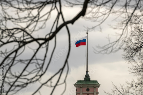 Flag of Russia with a view through the branches of a tree, develops on a spire against the background of clouds on a cloudy day