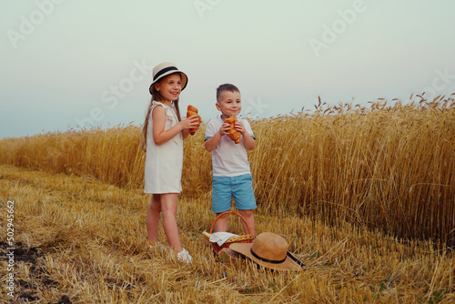 Little children wearing summer outfits standing by wheat field and eating croissants, basket and straw hat lying nearby. Happy siblings enjoying food in countryside at harvest season