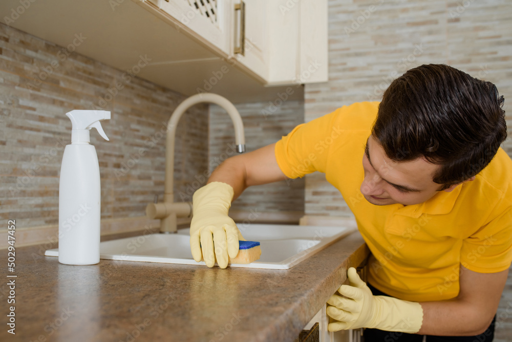 Close up young man in a yellow t-shirt and yellow gloves is cleaning the table in the kitchen using cleaning spray and sponge. Close up young man in a yellow t-shirt and yellow gloves is cleaning the table in the kitchen using cleaning spray and sponge.