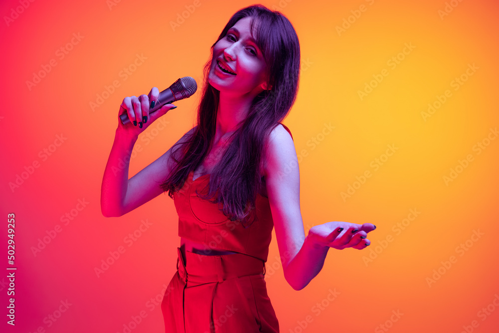 Naklejka premium Young excited girl, student with long hair singing isolated on orange background in neon light, filter. Concept of emotions, youth, beauty