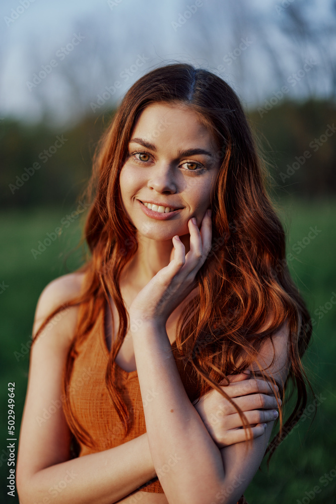 Fototapeta premium Portrait of a woman with a beautiful smile in summer green nature against the grass, looking out at the setting sun. The concept of health and beauty