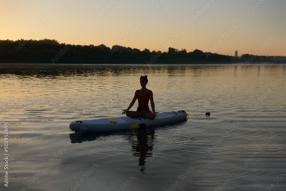 Silhouette of woman sitting and meditating on a paddle board during the fresh and warm summer morning