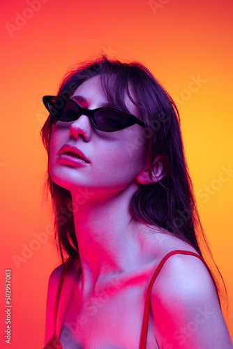 Stylish model in sunglasses. Young pretty girl with long glossy hair isolated over red-yellow background in neon light. Concept of emotions, art, beauty