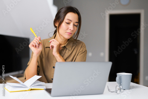 Smiling asian businesswoman taking notes sitting at the desk in modern office space. Young multiracial female student overwrites from the laptop screen into notebook photo