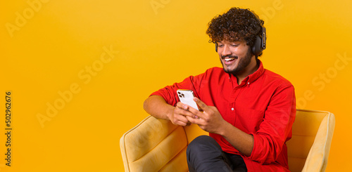 Studio portrait of young middle eastern guy in wireless headphones isolated on orange wall, looking at mobile phone, enjoying listening to favorite music soundtrack © Vadim Pastuh