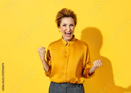 Overjoyed woman clenches fists with happiness, widely opened mouth as shouts loudly. Photo of positive elderly attractive woman in yellow shirt on yellow background © Romario Ien