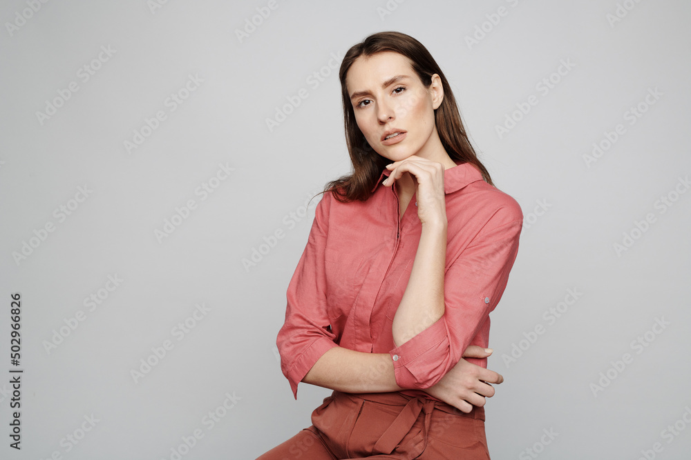 Portrait of content attractive young woman in pink shirt posing for camera and touching chin against white background Portrait of content attractive young woman in pink shirt posing for camera and touching chin against white background