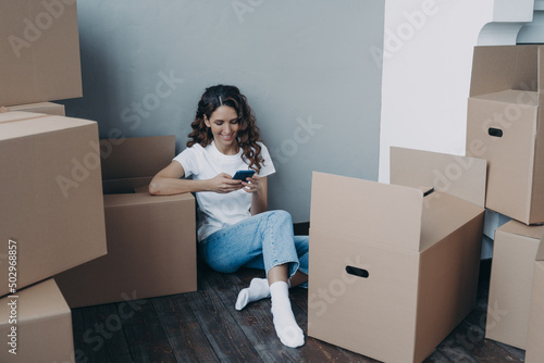Girl texting on mobile phone and sitting on the floor surrounded with cardboard packages and boxes.