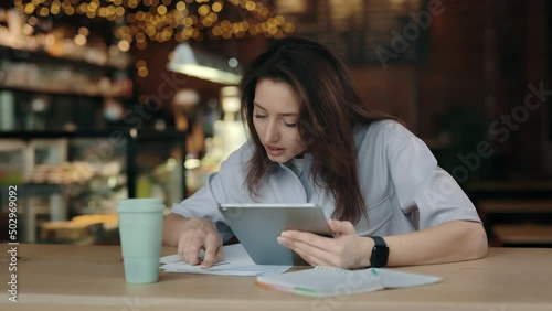 Beautiful caucasian brunette looking through some papers while sitting tafe with digital tablet hands. Young woman in casual attire spending leisure to for studying. photo