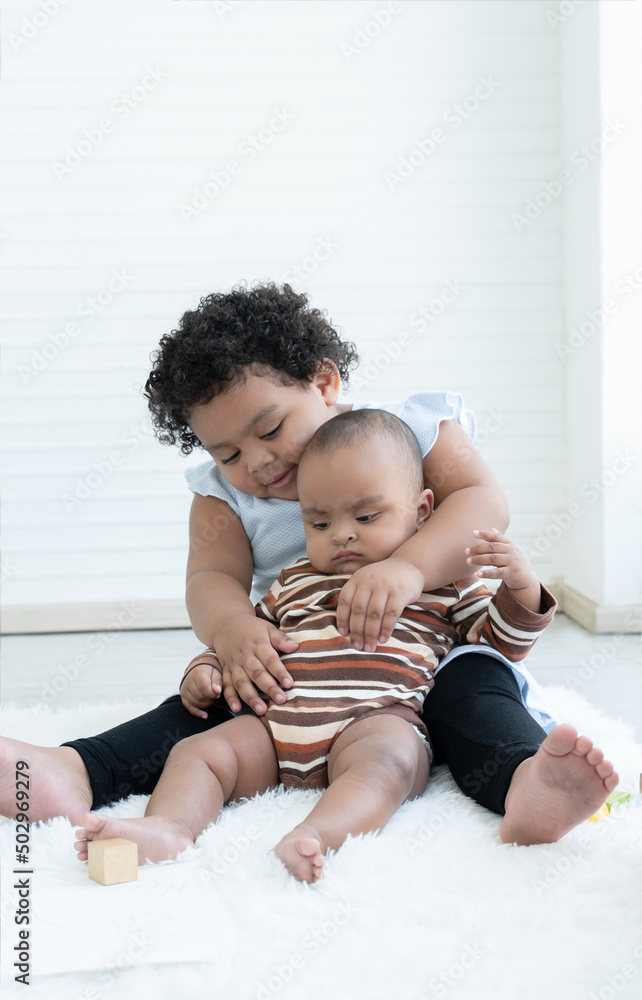 Happy Little cute African chubby kid girl hugging her newborn baby sister in arms while sitting playing together on floor at home. Siblings bond relationship concept. White background Happy Little cute African chubby kid girl hugging her newborn baby sister in arms while sitting playing together on floor at home. Siblings bond relationship concept. White background