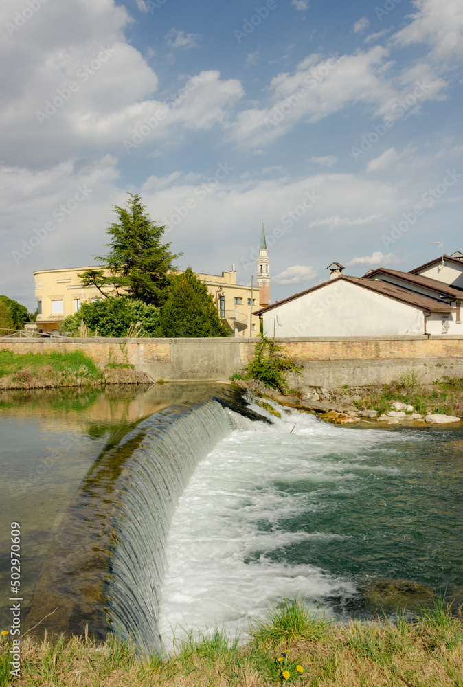 Italian rural landscape. Meschio river with a small artificial waterfall with the background of the houses and the sky with clouds. Vertical image. Cordignano, Treviso, Italy.
