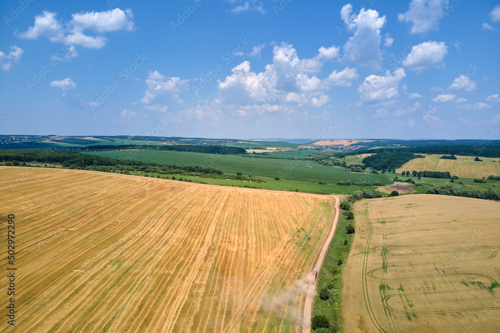 Obraz premium Aerial view of cargo truck driving on dirt road between agricultural wheat fields making lot of dust. Transportation of grain after being harvested by combine harvester during harvesting season