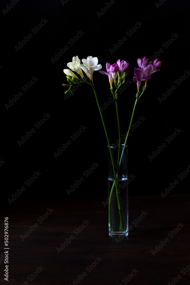 A bouquet of freesia stands in a transparent glass vase on a wooden table. Black background, low key, vertical orientation. A bouquet of freesia stands in a transparent glass vase on a wooden table. Black background, low key, vertical orientation.