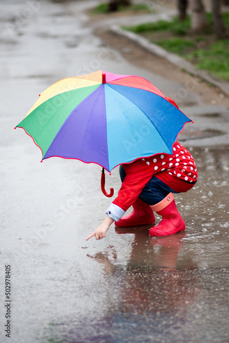 A cute little girl in a red cape, red boots and a white hat jump in puddles and has a fun.The girl has a rainbow umbrella in her hands. Happy childhood. Early spring. Emotions.