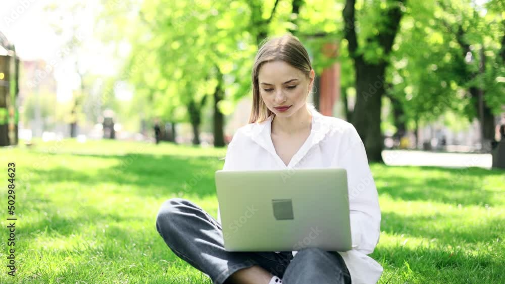 Attractive blond woman working at the laptop as sitting on grass in city park. Freelancer working in the park. Student study outside. Attractive blond woman working at the laptop as sitting on grass in city park. Freelancer working in the park. Student study outside.