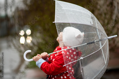 A cute little girl in a red cape, red boots and a white hat jump in puddles and has a fun.The girl has a transparent umbrella in her hands. Happy childhood. Early spring. Emotions.