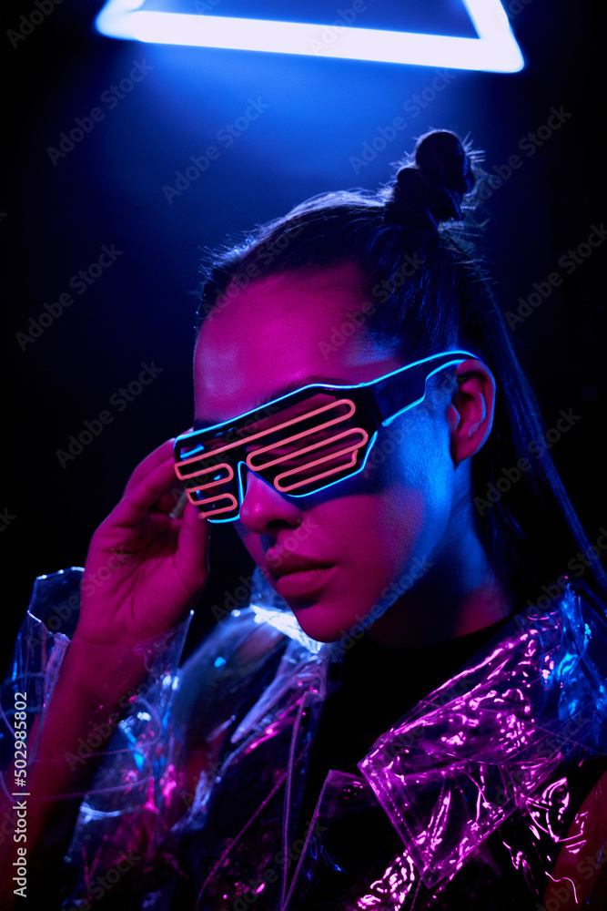 Portrait of ultramodern girl in transparent jacket adjusting flashing goggles in darkness