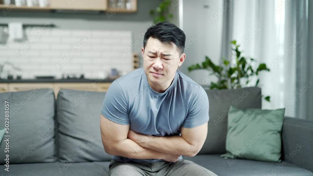 Asian man suffers from abdominal pain while sitting at home on the couch. stomach ache. diarrhea, gastritis problem, heartburn, severe stomach spasms Asian man suffers from abdominal pain while sitting at home on the couch. stomach ache. diarrhea, gastritis problem, heartburn, severe stomach spasms