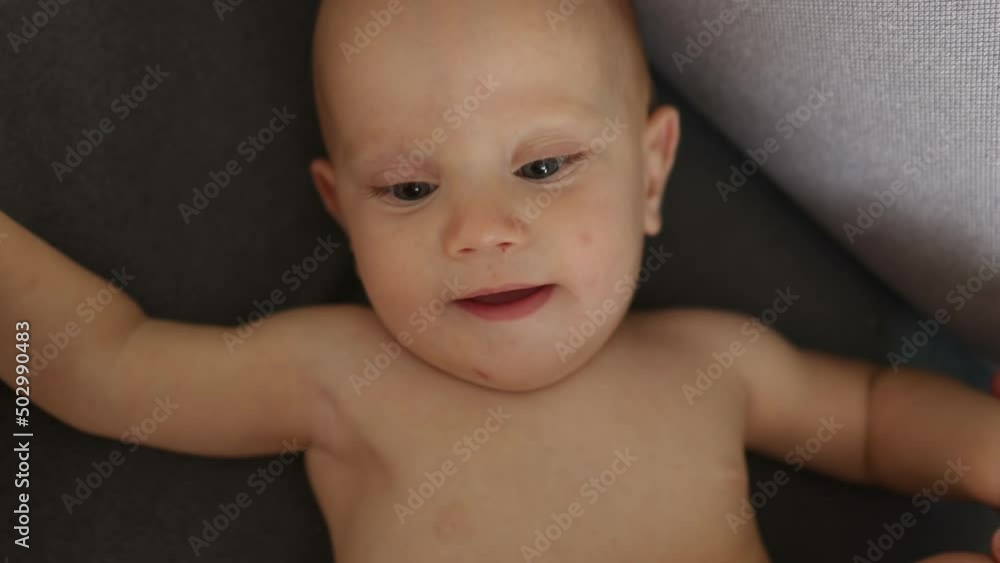 portrait of funny blue eyed naked baby infant smiling, laughing, looking at camera. little toddler kid laying on back on grey sofa, playing with mother's hands. happy caucasian child at home portrait of funny blue eyed naked baby infant smiling, laughing, looking at camera. little toddler kid laying on back on grey sofa, playing with mother's hands. happy caucasian child at home