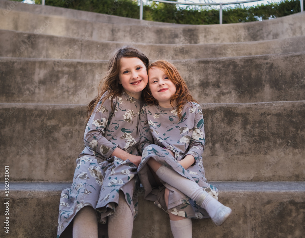 Portrait of two preteen sisters sitting in outdoor amphitheater smiling; girls wear similar dresses