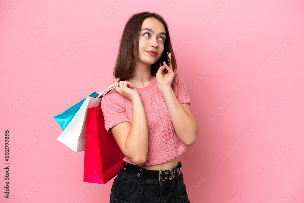 Young Ukrainian woman isolated on pink background holding shopping bags and calling a friend with her cell phone