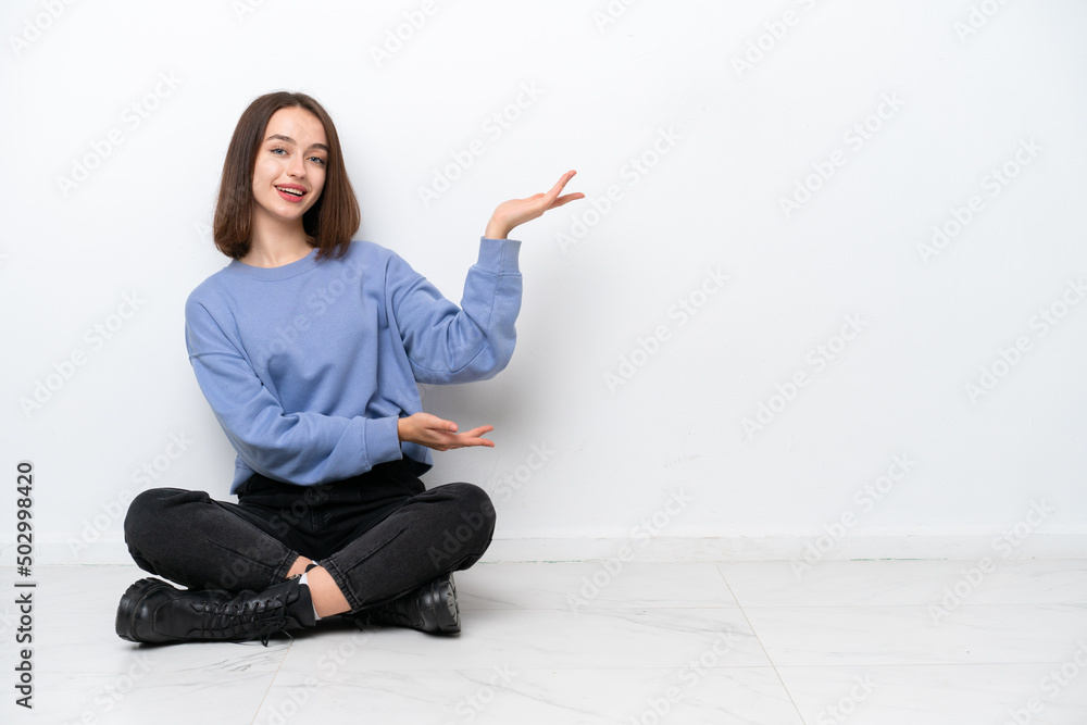Young Ukrainian woman sitting on the floor isolated on white background extending hands to the side for inviting to come Young Ukrainian woman sitting on the floor isolated on white background extending hands to the side for inviting to come