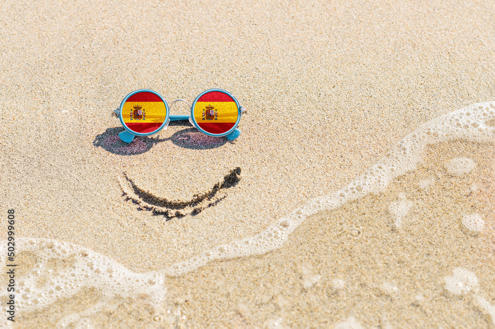 Naklejka premium A painted smile on the beach and sunglasses with the flag of Spain. The concept of a positive holiday in the resort of Spain.