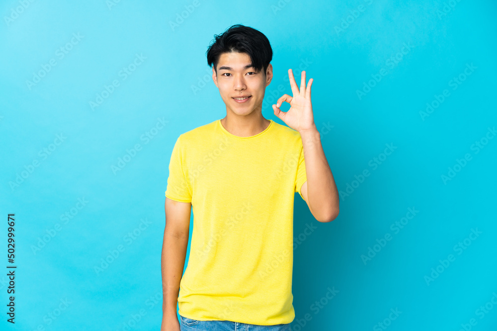 Young Chinese man isolated on blue background showing ok sign with fingers Young Chinese man isolated on blue background showing ok sign with fingers