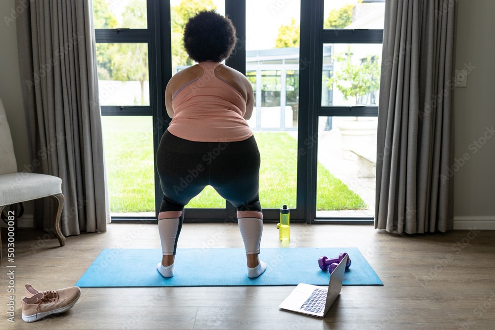 Rear view of african american mid adult woman doing squats on yoga mat at home Rear view of african american mid adult woman doing squats on yoga mat at home
