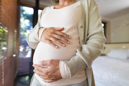 Mid section of caucasian pregnant woman touching her belly standing at home photo