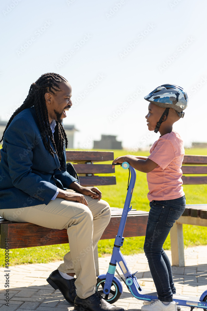 Happy african american father looking at son wearing helmet standing with push scooter on sunny day
