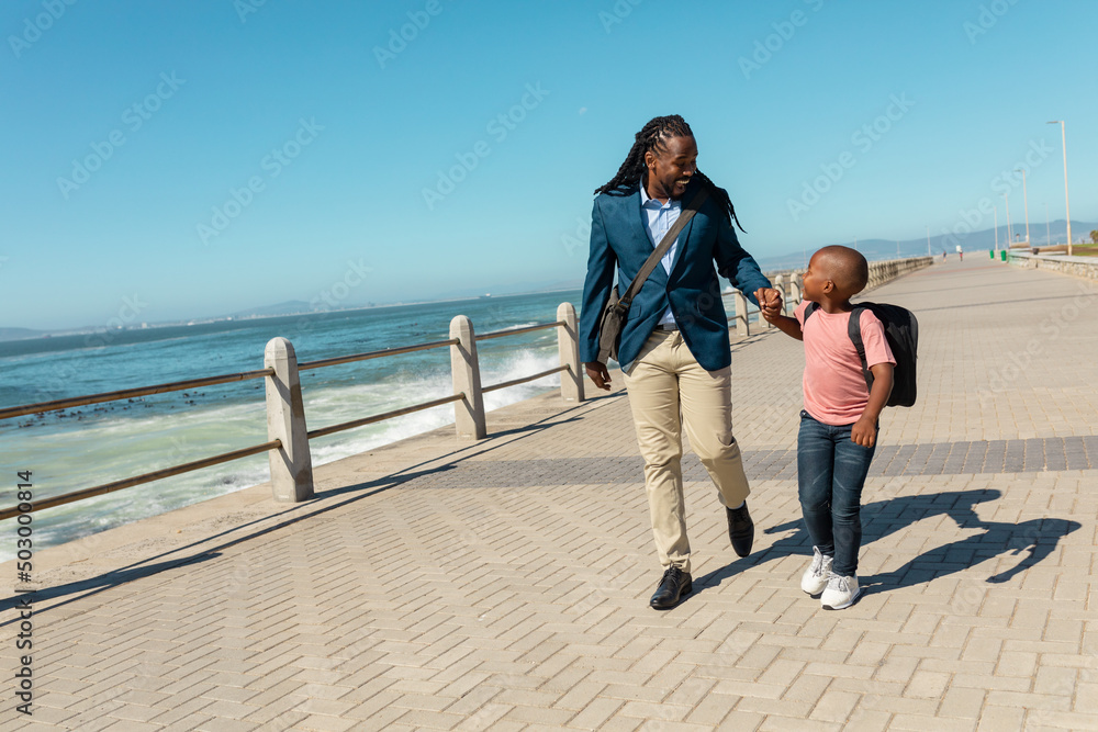 Happy african american father and son walking together on promenade against sky during sunny day Happy african american father and son walking together on promenade against sky during sunny day
