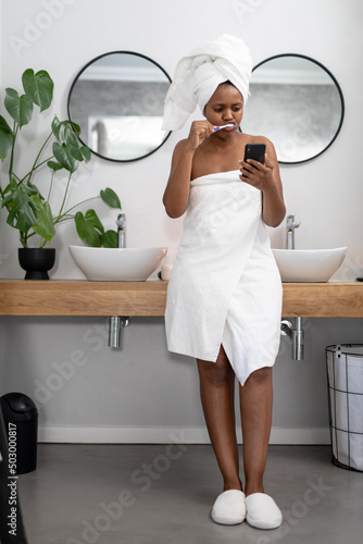 African american mid adult woman in towel using smart phone while brushing teeth in bathroom