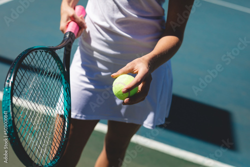 Midsection of young caucasian female tennis player standing with racket and ball at court
