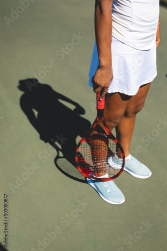 Low section of african american female athlete standing with tennis racket at court on sunny day