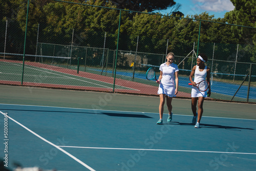 Smiling multiracial female players talking while walking at tennis court on sunny day