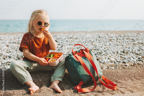 Child girl with lunchbox eating vegetables outdoor travel vacation healthy lifestyle vegan food picnic on beach hungry kid with lunch box snacks and backpack photo