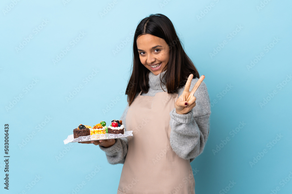 Pastry chef holding a big cake over isolated blue background smiling and showing victory sign. Pastry chef holding a big cake over isolated blue background smiling and showing victory sign.
