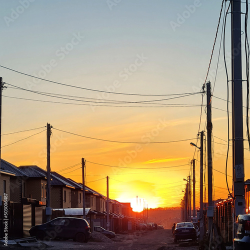 the sun's rays of an orange sunset in a Tatar village against the background of houses, electricity poles, wires and parked cars