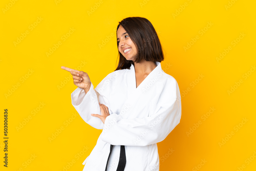 Young woman doing karate isolated on yellow background pointing finger to the side and presenting a product Young woman doing karate isolated on yellow background pointing finger to the side and presenting a product