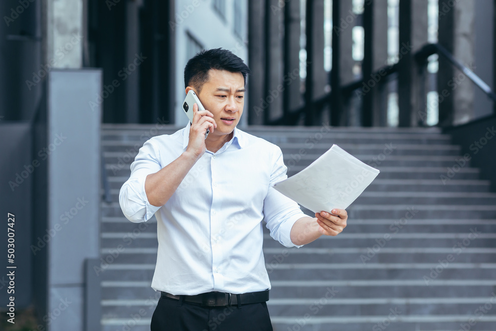 Frustrated insurance agent with documents talking on the phone outside the office, depressed Asian businessman Frustrated insurance agent with documents talking on the phone outside the office, depressed Asian businessman