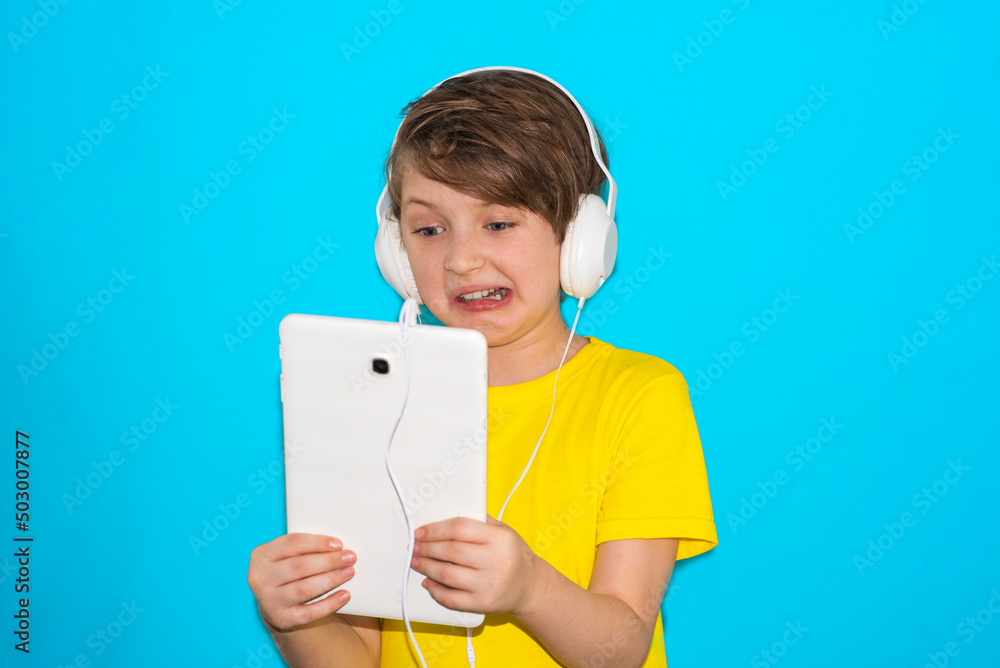 the boy communicates on the Internet. the baby is shocked. schoolboy in headphones holds a tablet in his hands. blue background
the boy communicates on the Internet. the baby is shocked. schoolboy in headphones holds a tablet in his hands. blue background