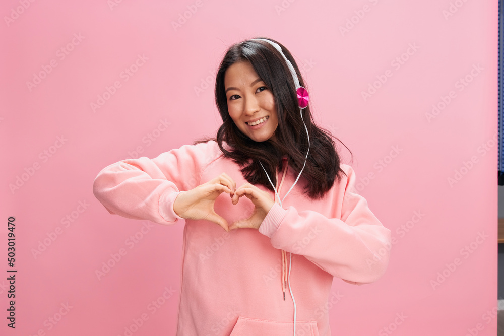 Friendly overjoyed Asian young lady in pink hoodie sweatshirt with cute headphones show heart sign gesture posing isolated on pink studio background. Good offer. Sound streaming platform ad concept
