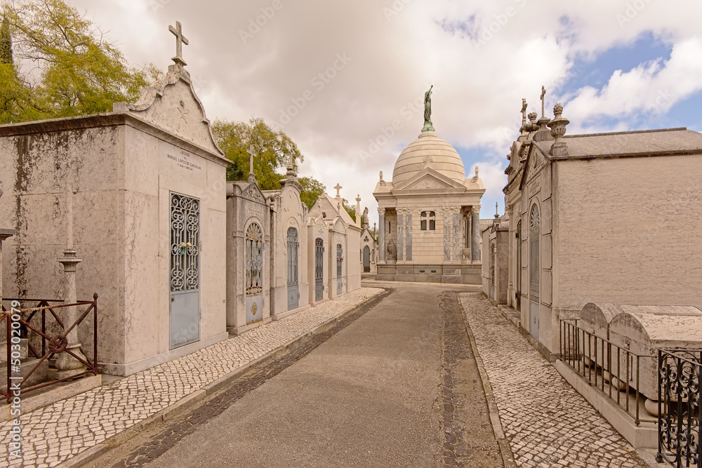 Rows of graves and ornate grave chapel with cupola with bronze statue of mourning woman, holding her arms up to the sky in Alto de Sao Joao cemetery, Lisbon, Portugal 