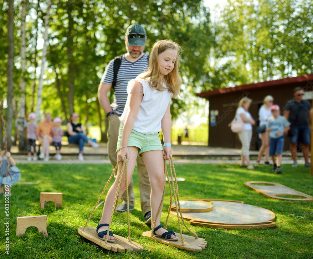 Naklejka premium Little girl trying to walk on giant wooden feet in the park. Child playing funny outdoor game on sunny summer day. Family leisure for kids and parents.