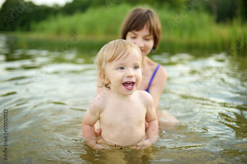 Cute toddler boy and his big teenage sister playing by a river on hot summer day. Adorable child having fun outdoors during summer vacations.
